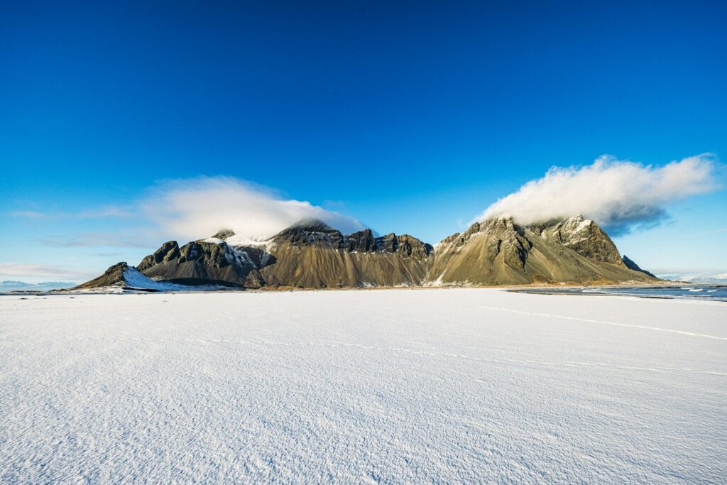 Vast snowfield before rugged mountains with clouds under blue sky.